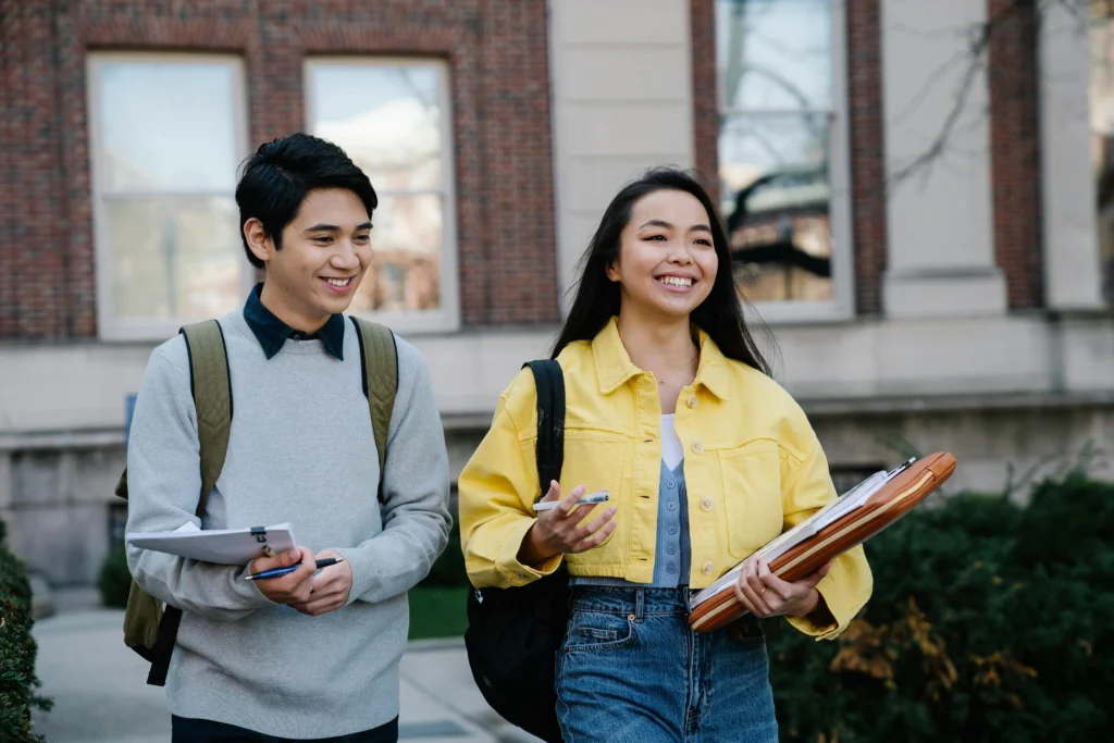 Two students stroll across campus, carrying folders, as they discuss their studies in a lively outdoor setting.