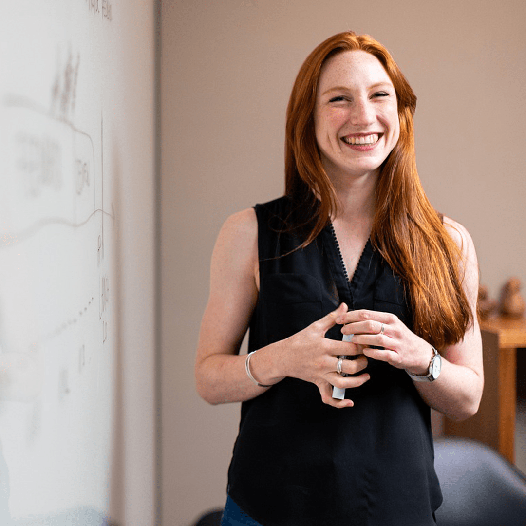 A woman with long red hair, wearing a sleeveless black top, stands and smiles near a whiteboard with diagrams in a modern office setting.