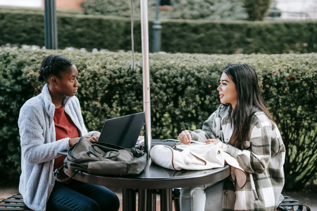 Two women engaged in conversation while working on laptops at a table.