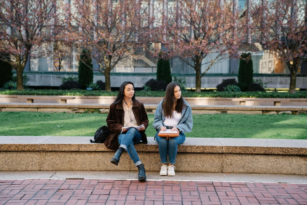 Two young women sit on a bench in front of a building, engaged in conversation and enjoying their time together.