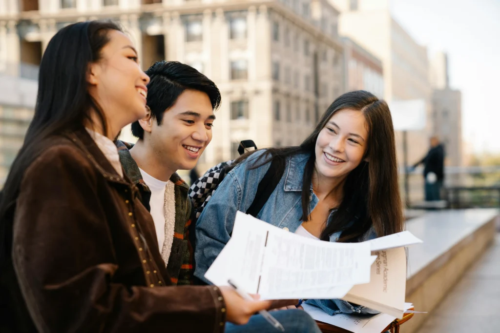Three students sit outdoors, smiling and holding papers, appearing to study or discuss coursework together in an urban campus setting.